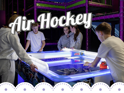 Im Bild sieht man einen Blau-Weißen Air Hockey Tisch. Zwei Jungen spielen gegeneinander und 3 Mädchen stehen daneben und schauen zu.