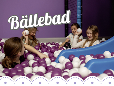 Das Bällebad besteht aus violetten und weißen Bällen. 4 Mädchen sitzen darin und spielen mit den Bällen. Rechts ragt eine kleine blaue Rutsche ins Bild hinein.