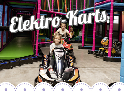 Im Bild sieht man eine Elektro-Kartbahn. 2 E-Karts, die von Kindern gefahren werden, fahren direkt auf die Kamera zu. Die Strecke verläuft unter dem Klettergerüst durch.